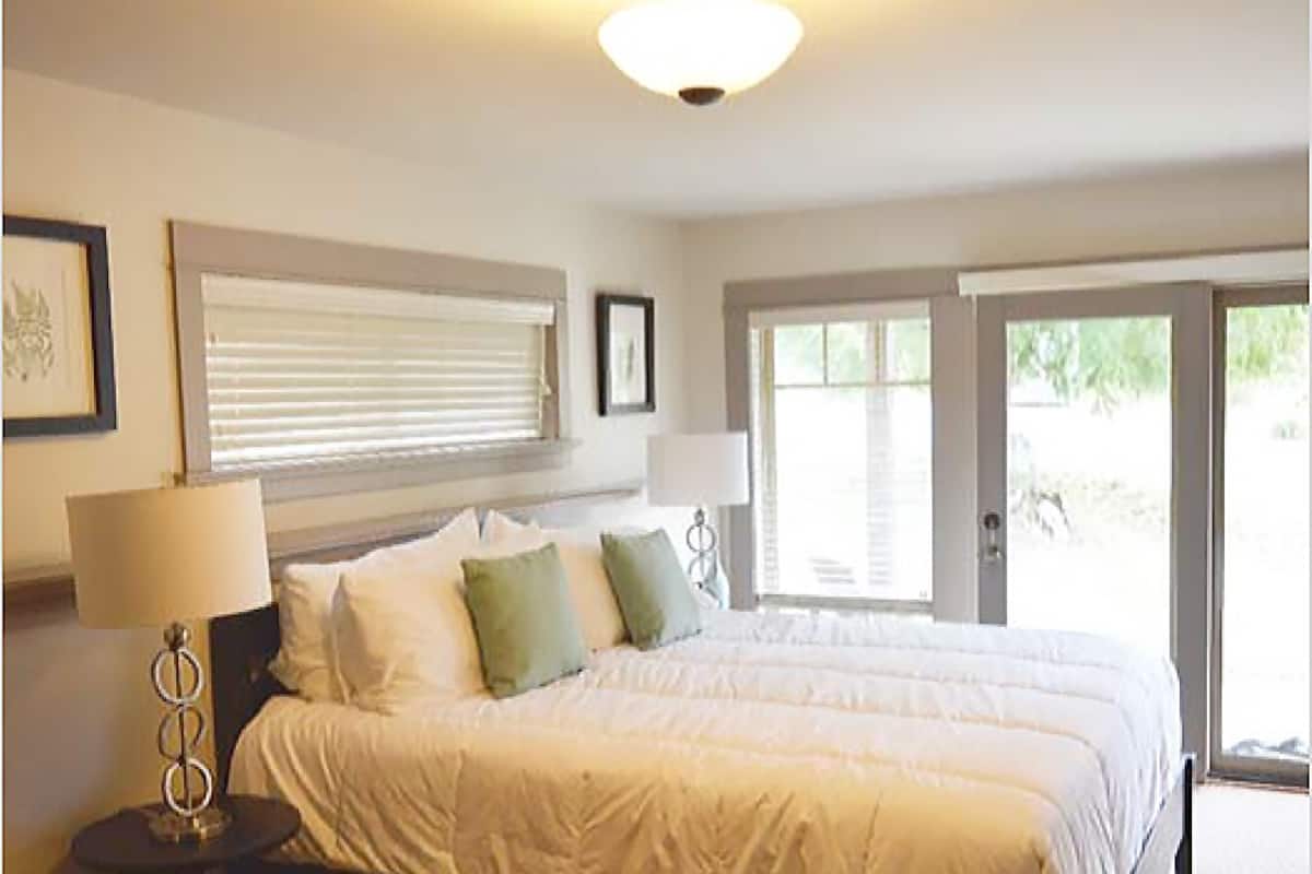 A bedroom with a large bed, a bedside table with a lamp, and a window with blinds.