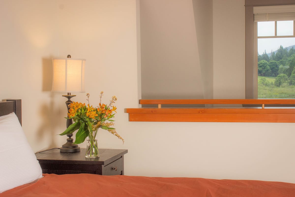 A bedroom with a bed, a nightstand with a lamp and flowers, a window with a view of greenery, and a wooden shelf above the window.