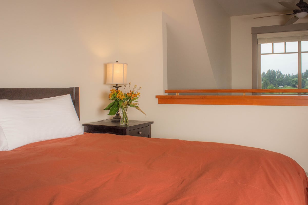 A bedroom with a bed, nightstand, lamp, flowers, and a window with a view.