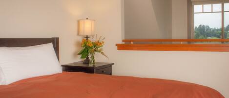 A bedroom with a bed, nightstand, lamp, flowers, and a window with a view.
