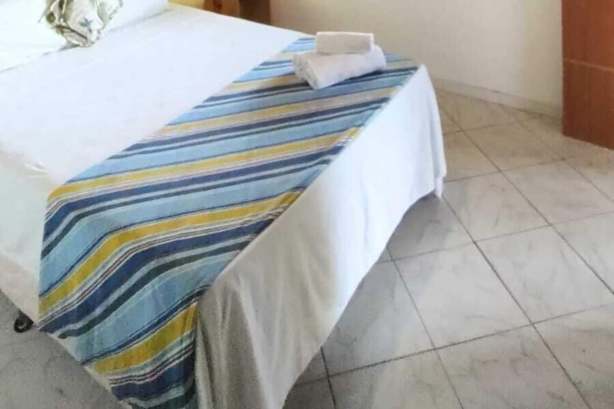 A hotel room with a wooden ceiling, a bed with a striped bedspread, an air conditioning unit, a window with shutters, and a door.