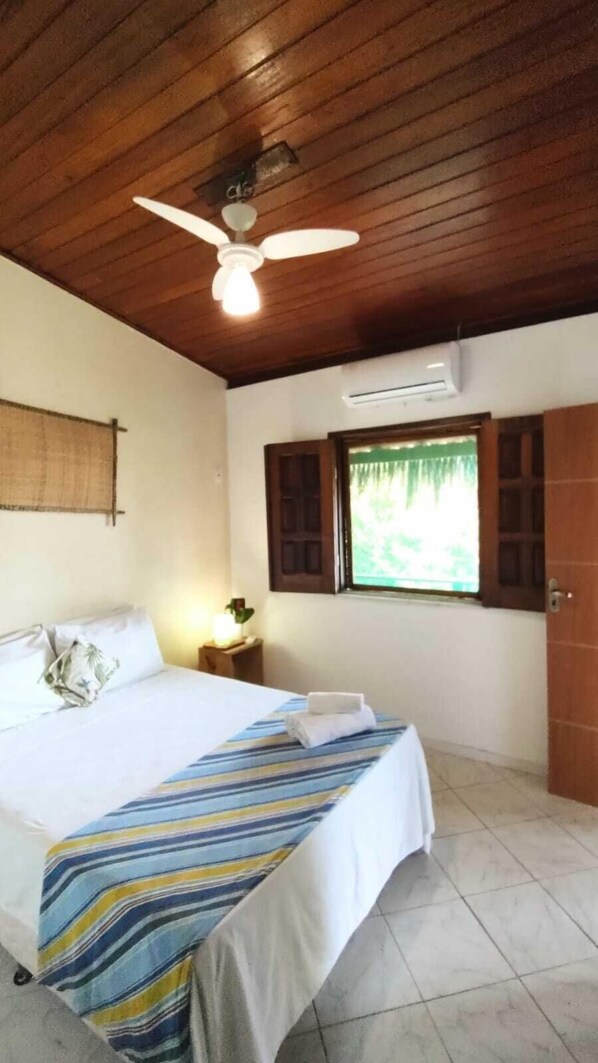 Quarto de hotel com teto de madeira, cama com colcha listrada, ar condicionado, janela com persianas e uma porta.