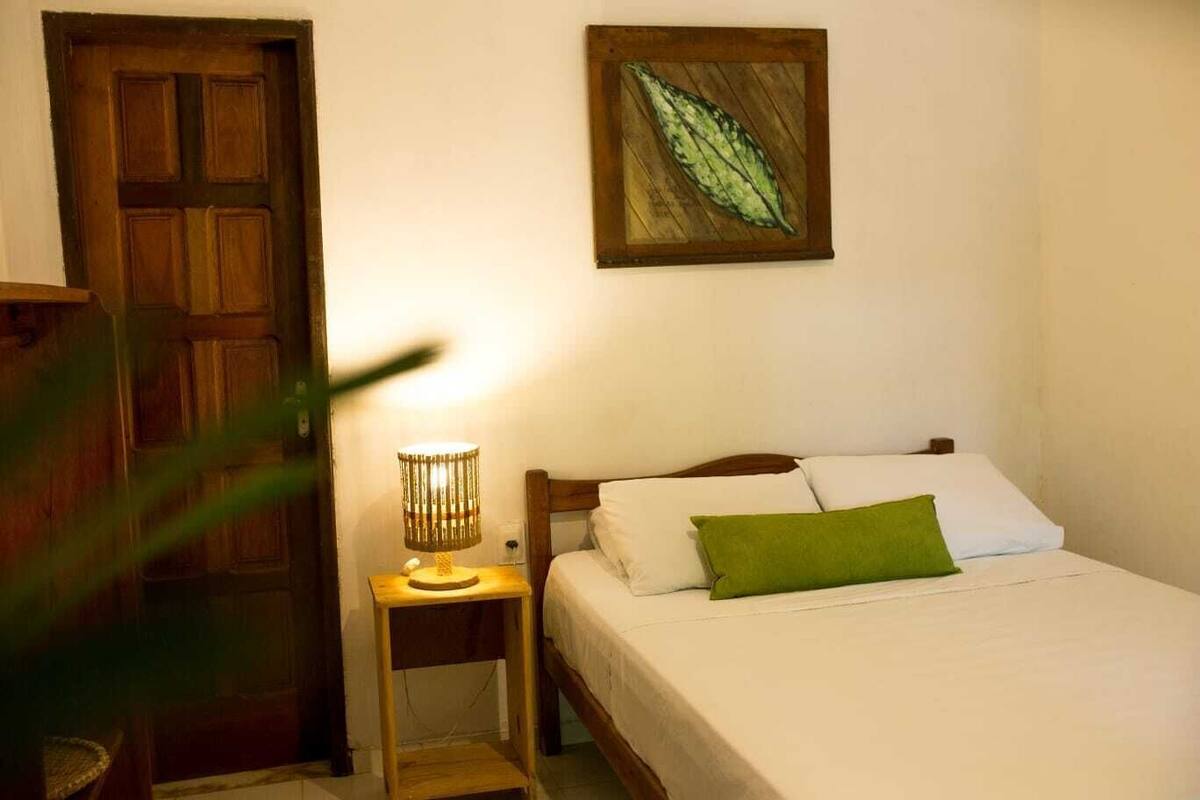 A bedroom with a wooden door, a bedside lamp, a bed with white bedding and a green pillow, and a leaf-patterned artwork on the wall.