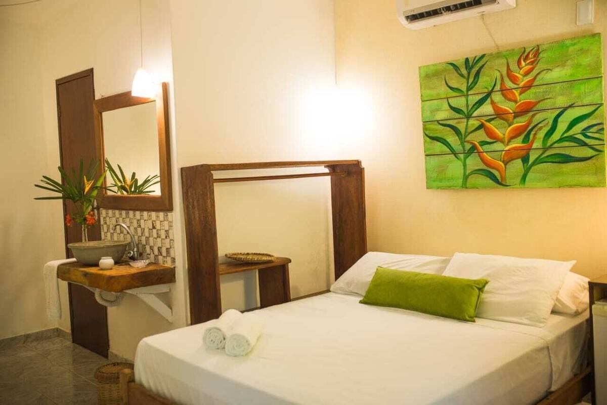 A hotel room with a bed, a wooden headboard, a painting on the wall, a sink with a mirror, and a potted plant.