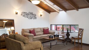 Ein Wohnzimmer mit einem beigen Sofa, einem Holztisch und einem Holzstuhl. Ein Fenster bietet Blick auf ein rotes Gebäude.