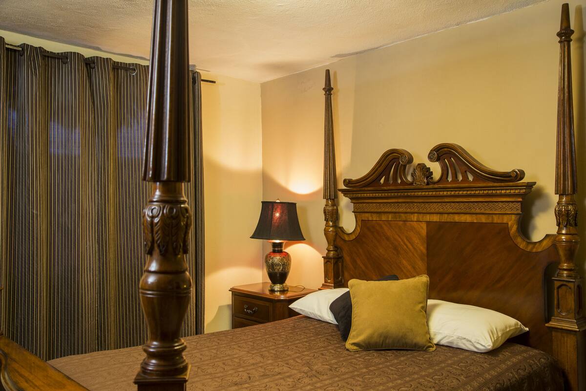 Een slaapkamer met een hemelbed, een nachtkastje met een lamp en een raam met gordijnen.