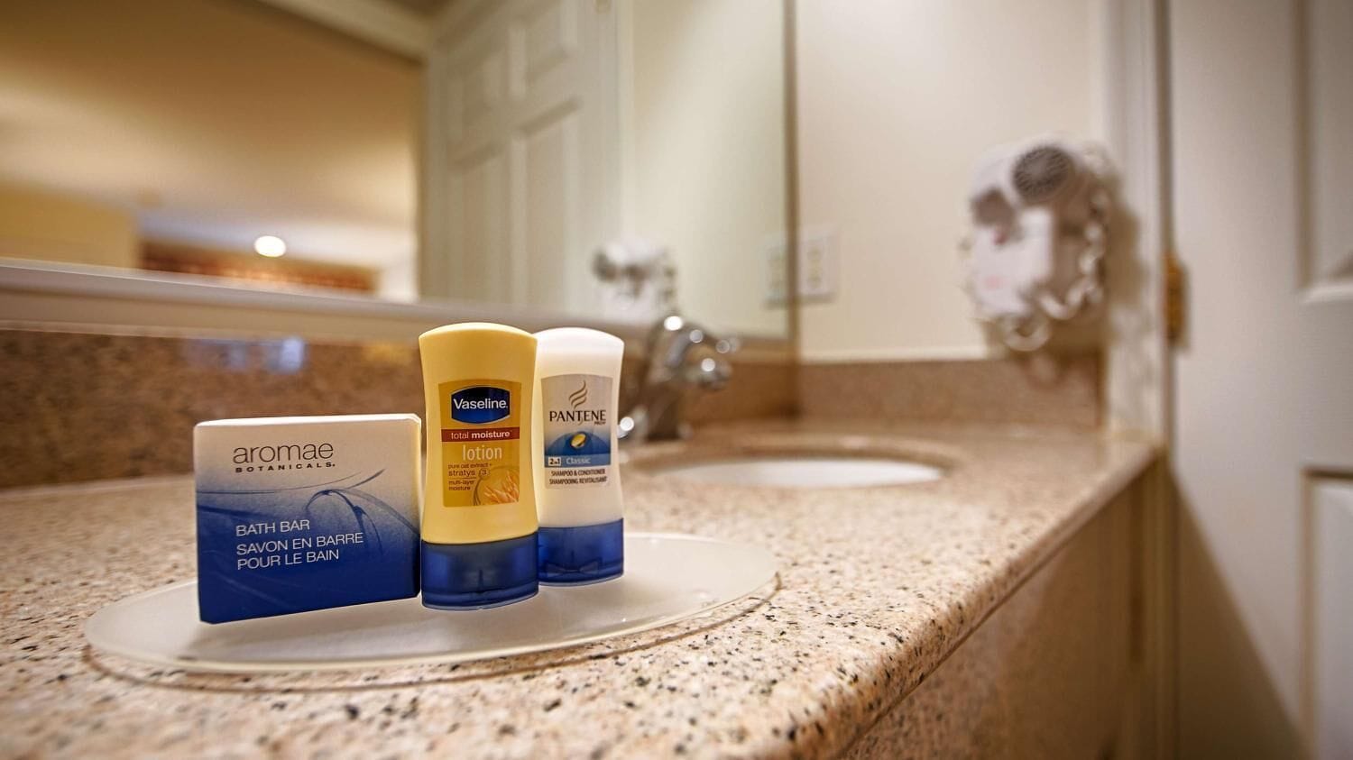 A bathroom counter with a mirror, a sink, and three toiletries: a bath bar, a lotion, and a shampoo.