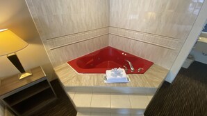 A corner bathroom with a red bathtub, a white towel, and a lamp on a wooden nightstand. - Garden State Inn (Absecon)