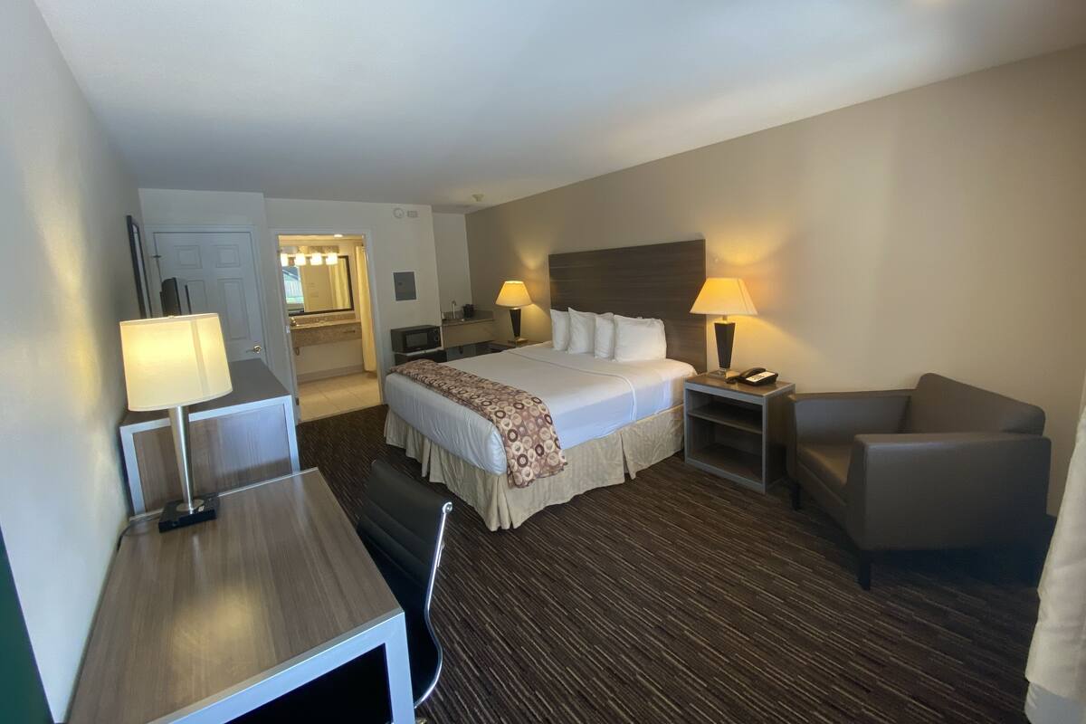 A hotel room with a bed, two bedside tables, a desk, a chair, and a sofa. There is a bathroom visible in the background.