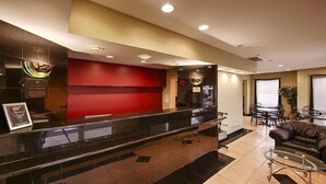 A hotel lobby with a reception desk, a red accent wall, a seating area with a leather couch and a glass table, and a dining area with chairs and a table. - Garden State Inn (Absecon)