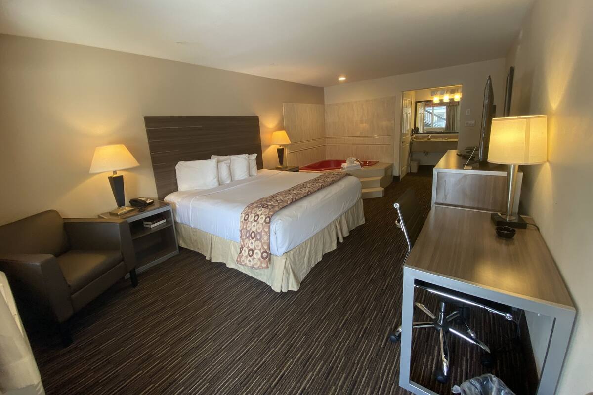 A hotel room with a large bed, bedside tables with lamps, a desk with a chair, a mirror, and a bathroom visible through an open door.