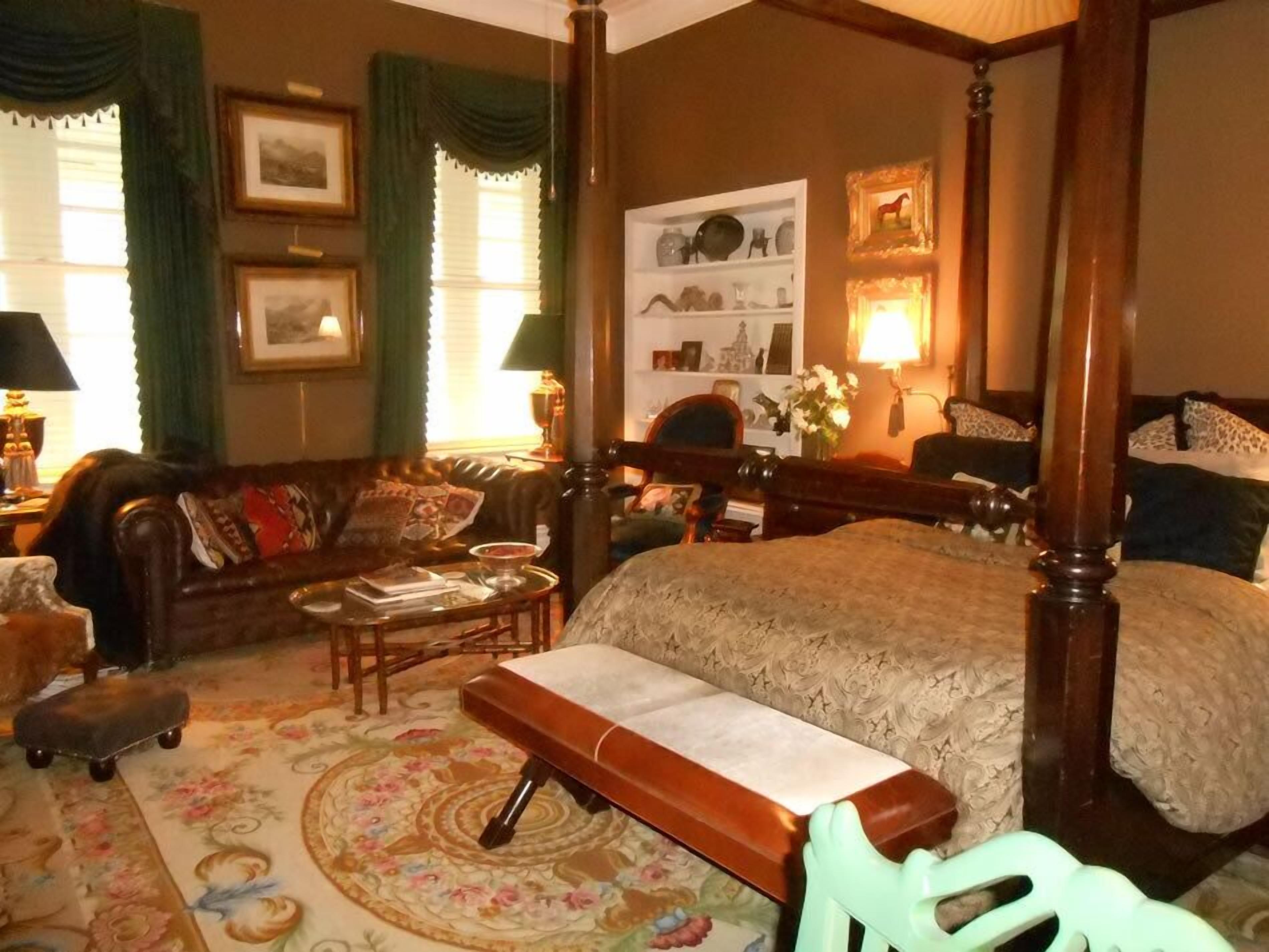 A traditional bedroom with a four-poster bed, a sitting area with a sofa and armchairs, a small table, and a decorative rug.