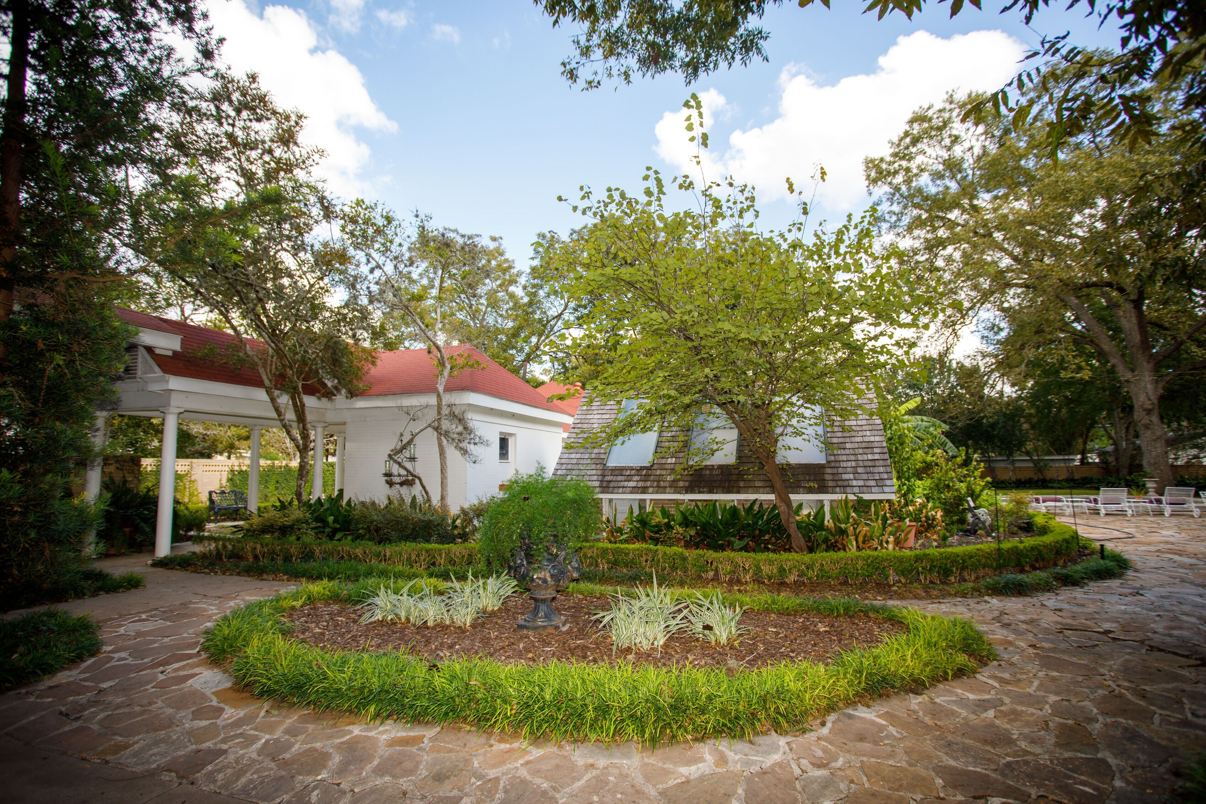 A courtyard with a stone pathway, a white building with a red roof, and a variety of plants and trees.