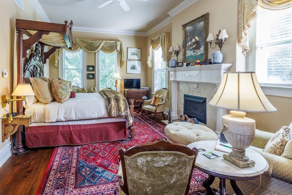 A spacious bedroom with a large bed, a fireplace, a ceiling fan, and a red patterned rug.