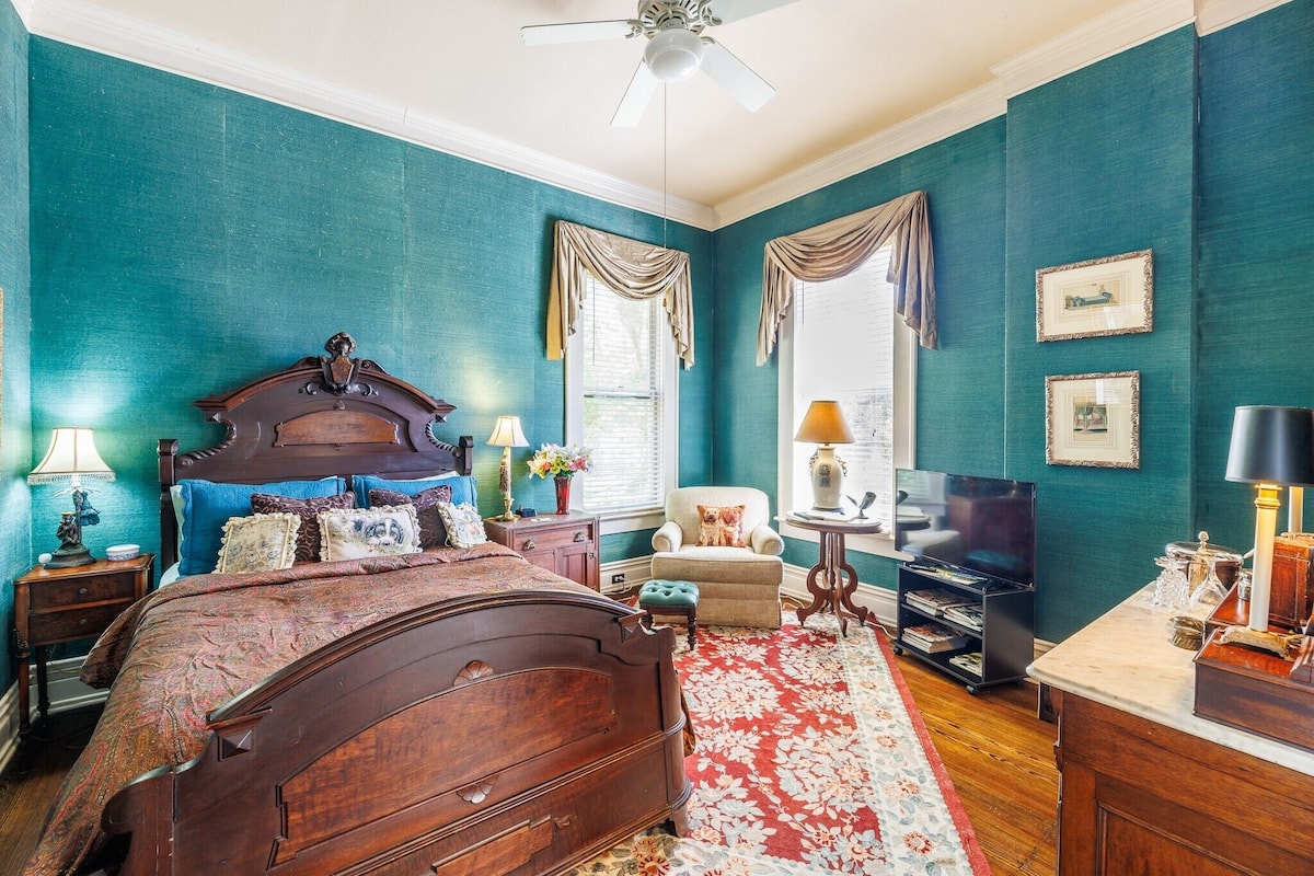 A bedroom with teal walls, a wooden bed, a red floral rug, a TV, and a chair.