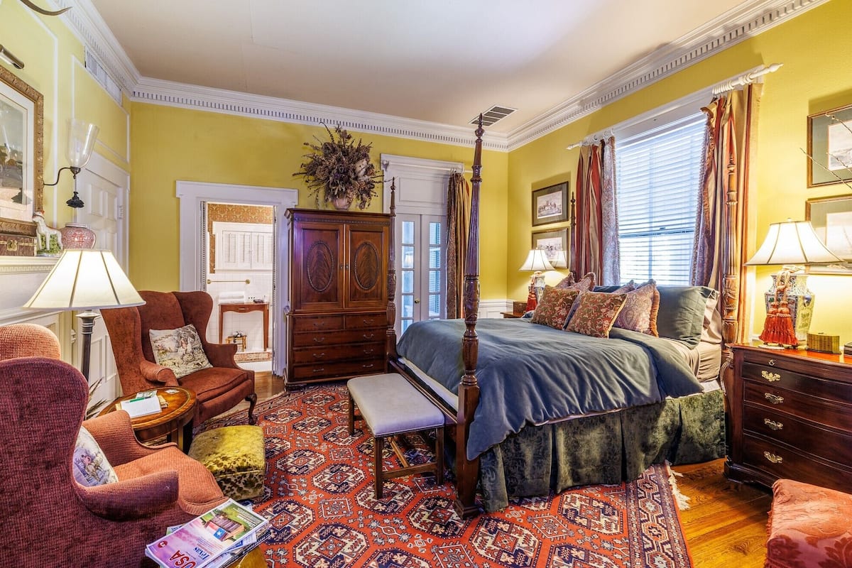 A traditional bedroom with a four-poster bed, a wooden wardrobe, a bench, and a patterned rug.