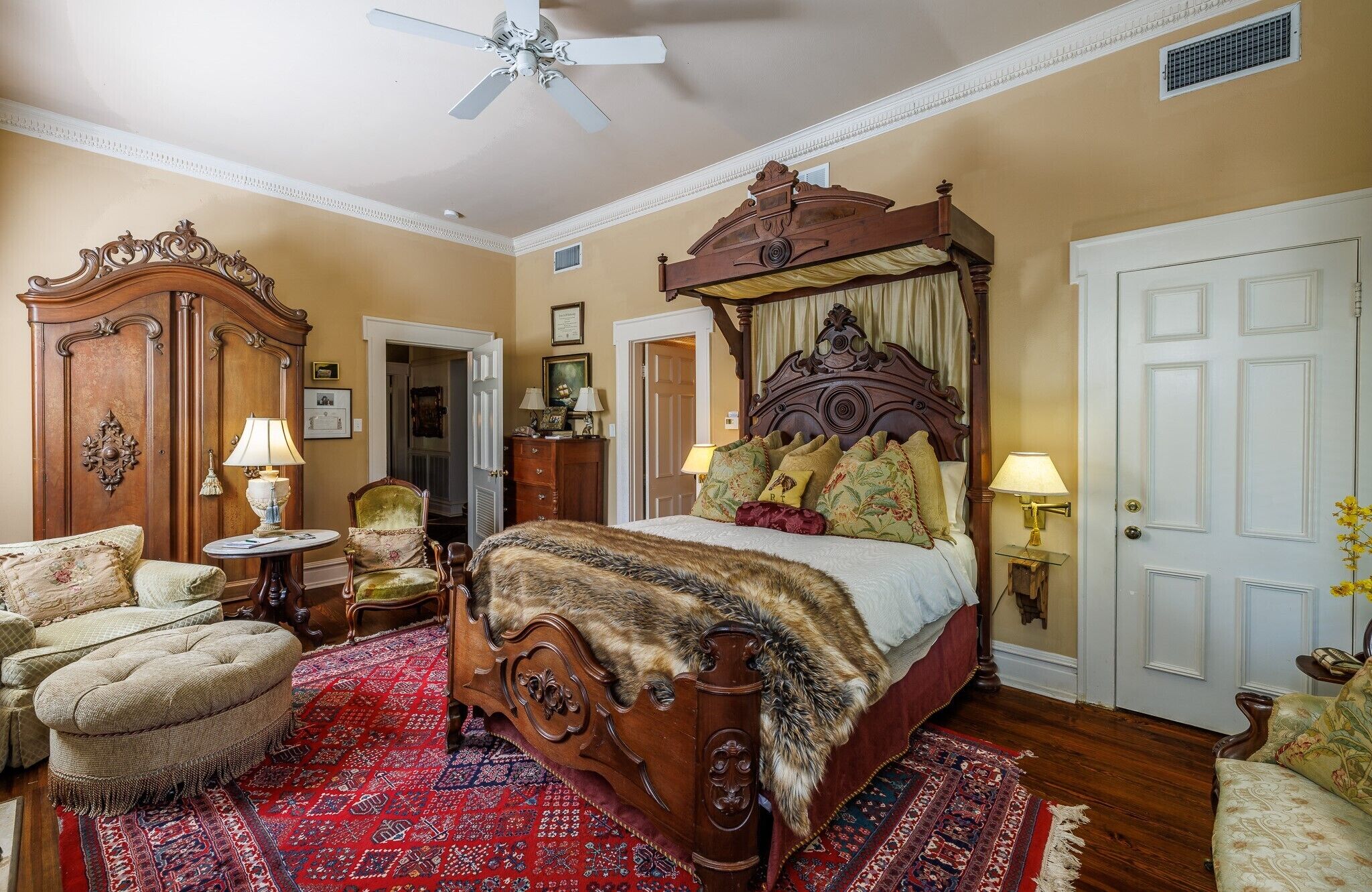 A traditional bedroom with a four-poster bed, a wooden wardrobe, a round table with a lamp, and a patterned rug.