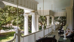 A covered porch with white columns and railings, wicker furniture, and a view of trees and a street. - Belle Oaks Inn (Gonzales)