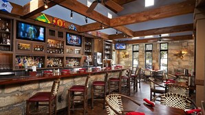 A bar area with wooden beams, stone walls, and multiple televisions. There are bar stools and tables with red cushions. - Hilton Houston Westchase (Houston)