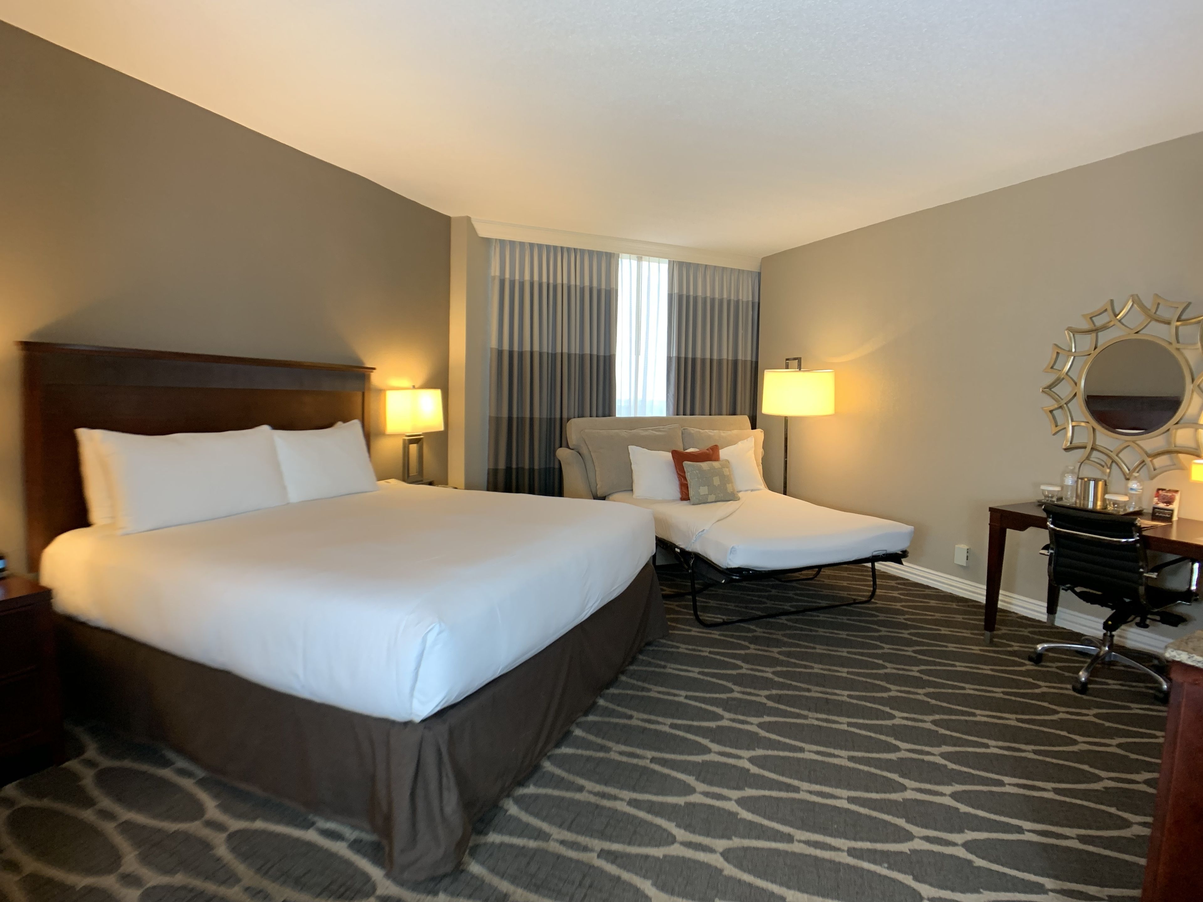 A hotel room with a large bed, a desk, and a chair. There are two lamps on the desk and a mirror on the wall.