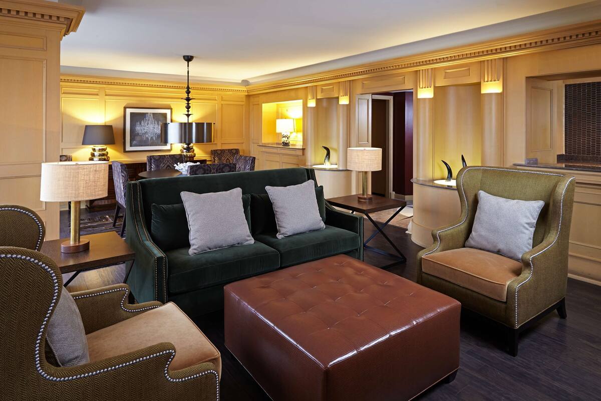 A spacious living room with a green sofa, a brown ottoman, and two armchairs. There is a television, a chandelier, and a dining area in the background.