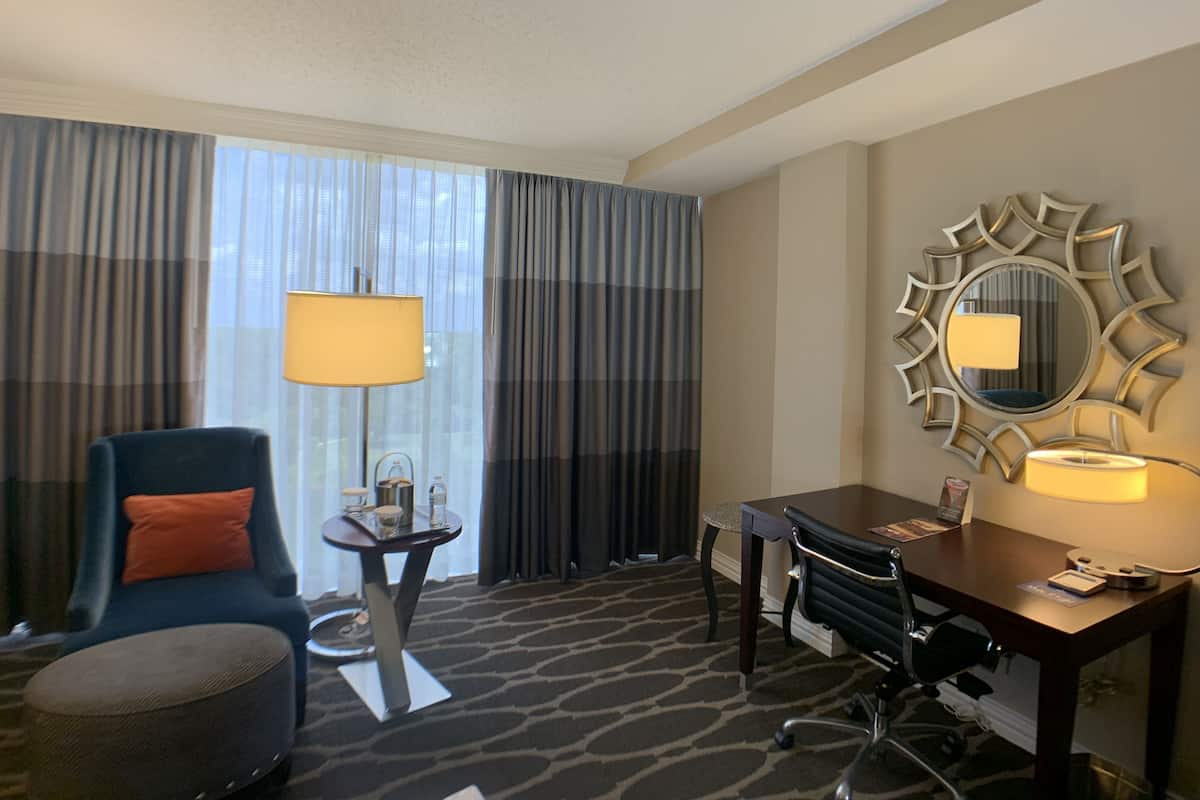 A hotel room with a large window, a desk with a chair, a round mirror, and a small table with a lamp.
