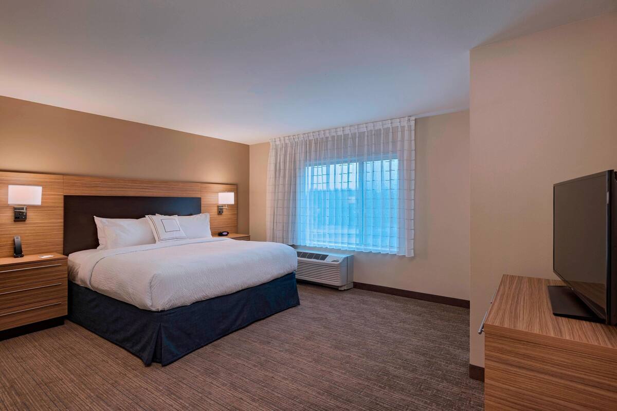 A hotel room with a large bed, bedside tables with lamps, a television, and a window with sheer curtains.