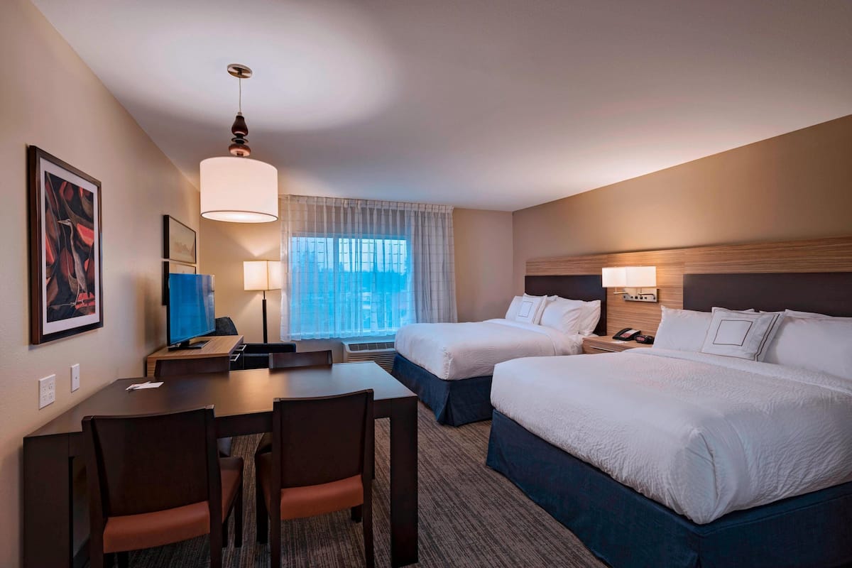 A hotel room with two beds, a desk, chairs, a TV, and a large window with curtains.