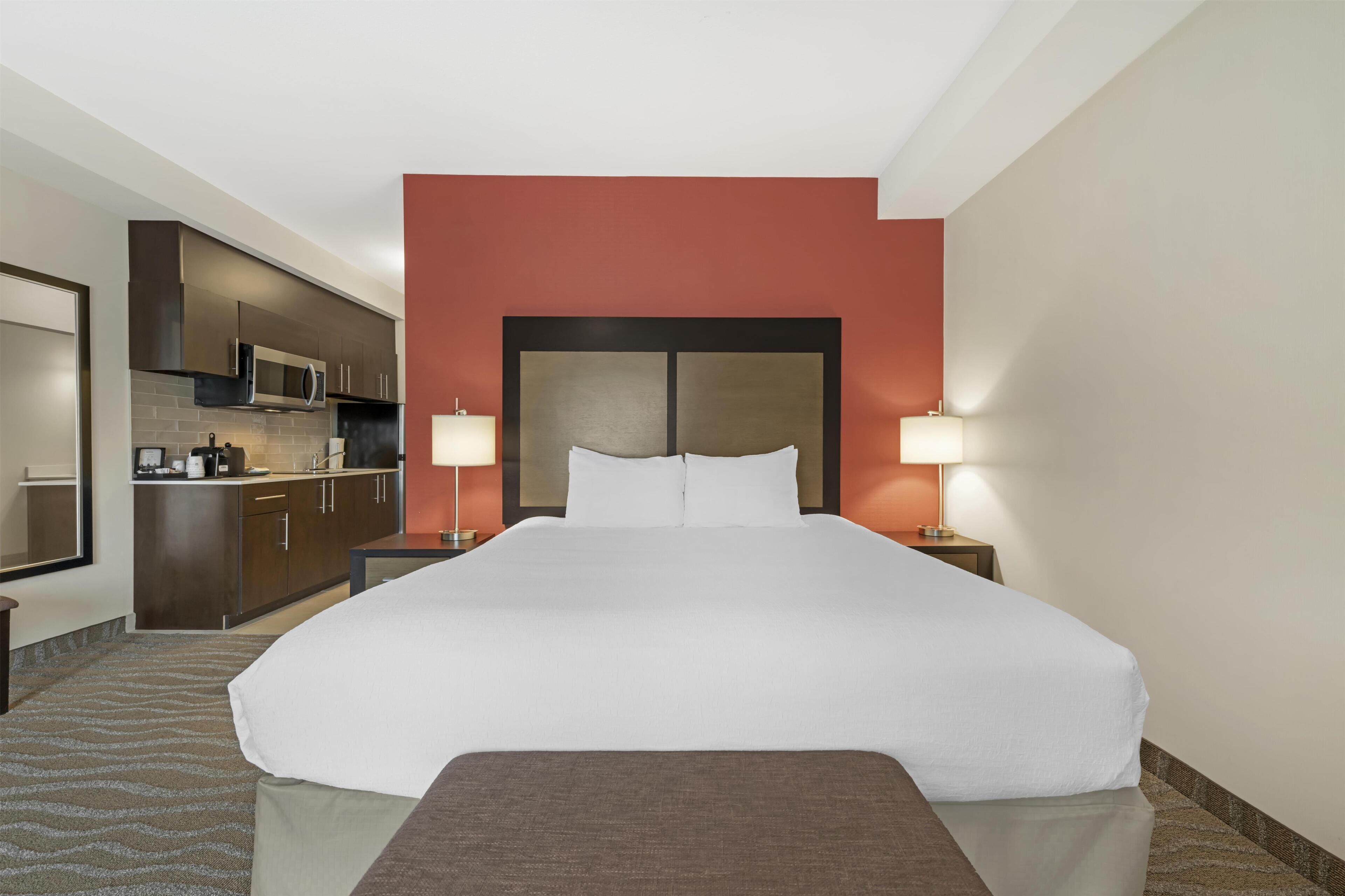 A hotel room with a large bed, a kitchenette, and two bedside lamps.