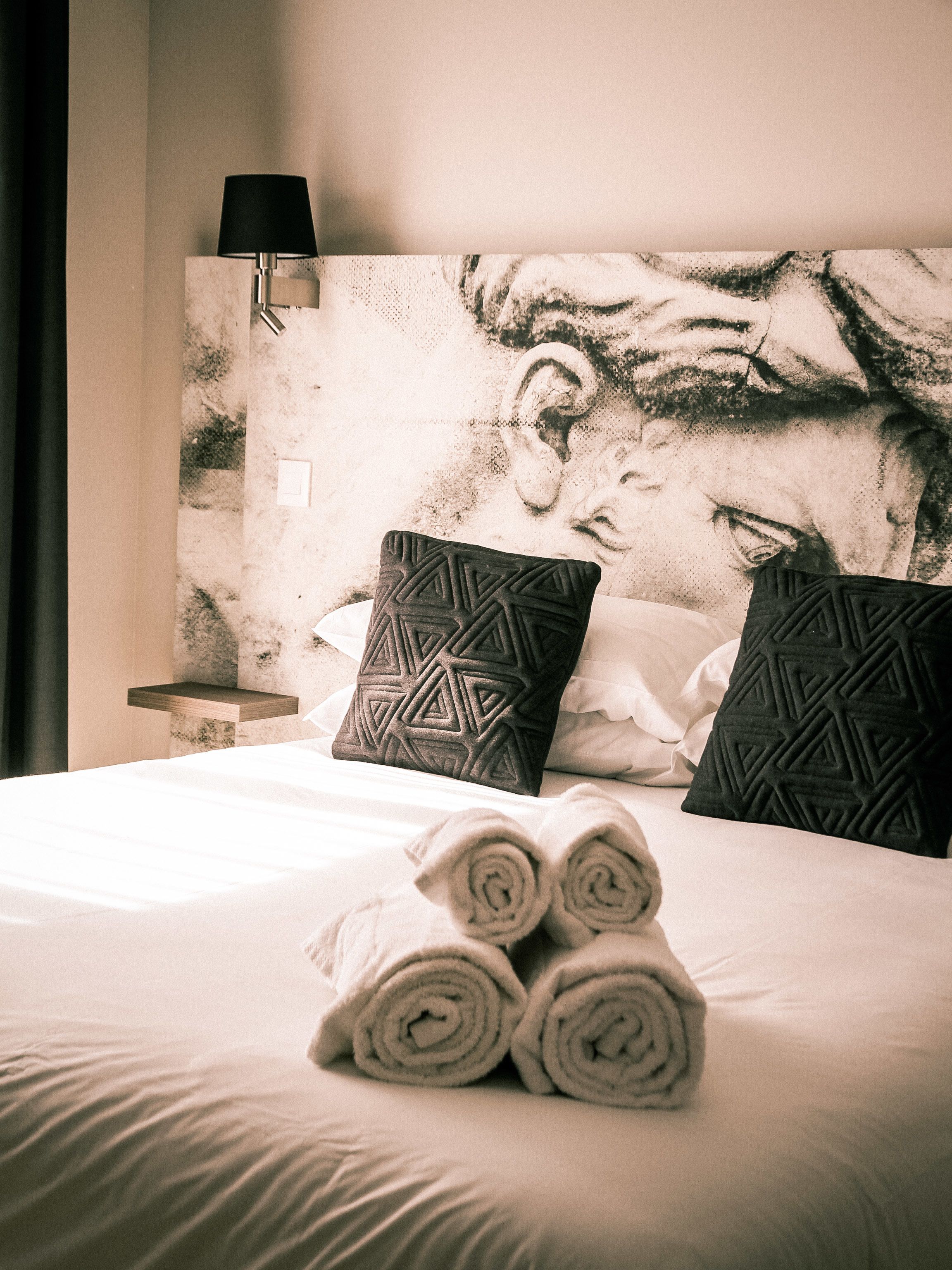Una cama bien tendida con toallas enrolladas, dos almohadas estampadas, una lámpara de pared y una vista parcial de un cuadro.