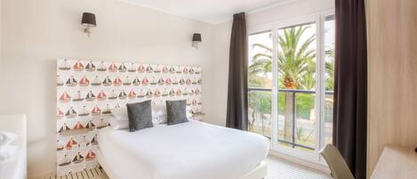 Habitación de hotel con una cama grande, un cabecero con estampado de velero, dos lámparas fijadas a la pared, vista a palmeras a través de un ventanal amplio, y suelo de madera.