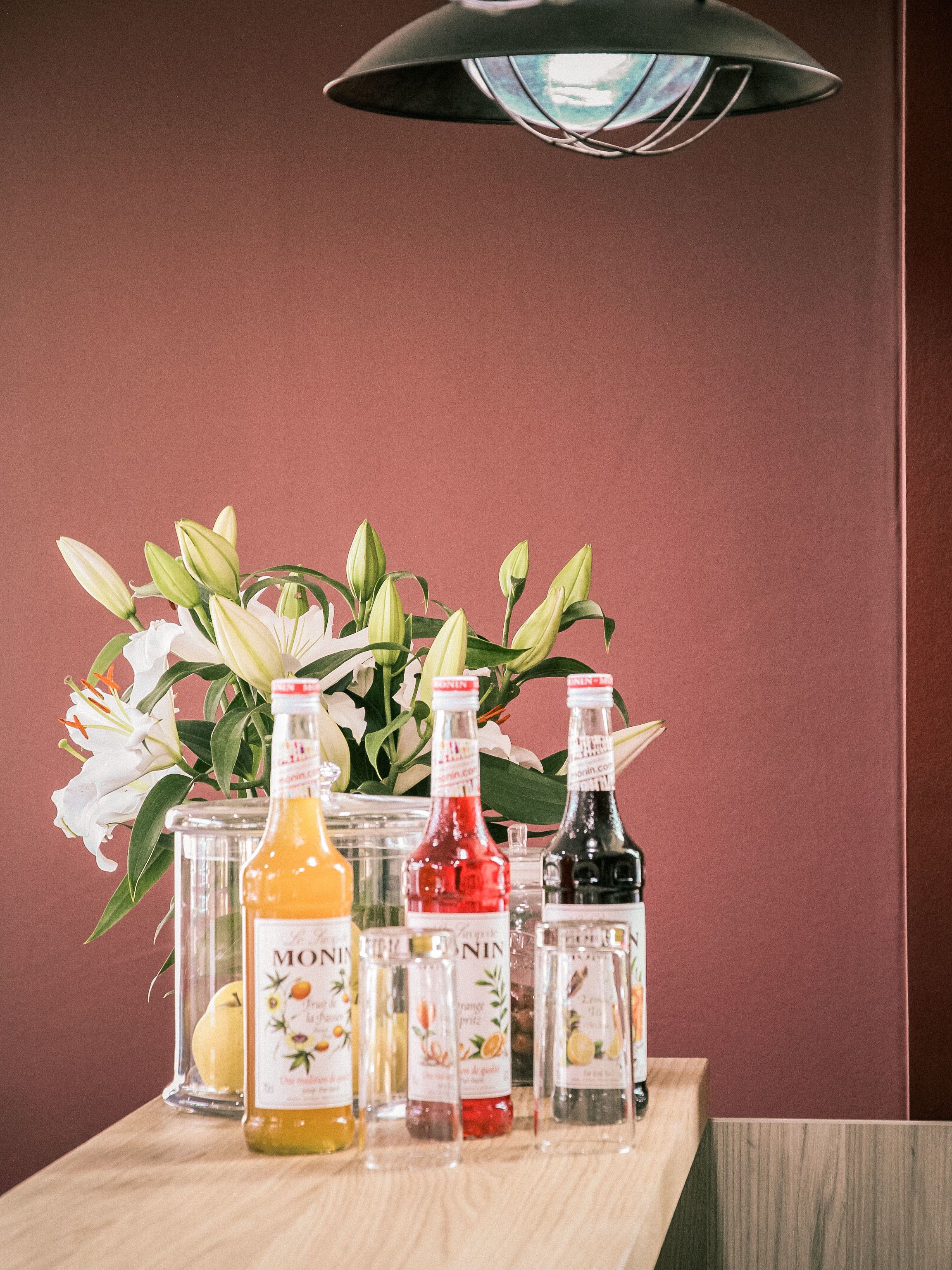 Una mesa con botellas de sirope Monin y un jarrón con lirios blancos frente a una pared color burdeos.