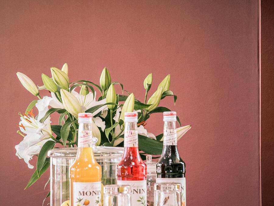 Una mesa con botellas de sirope Monin y un jarrón con lirios blancos frente a una pared color burdeos.