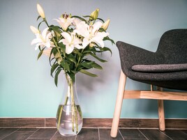 Eine Vase mit weißen Blumen und ein Holzstuhl vor einer türkisfarbenen Wand.
