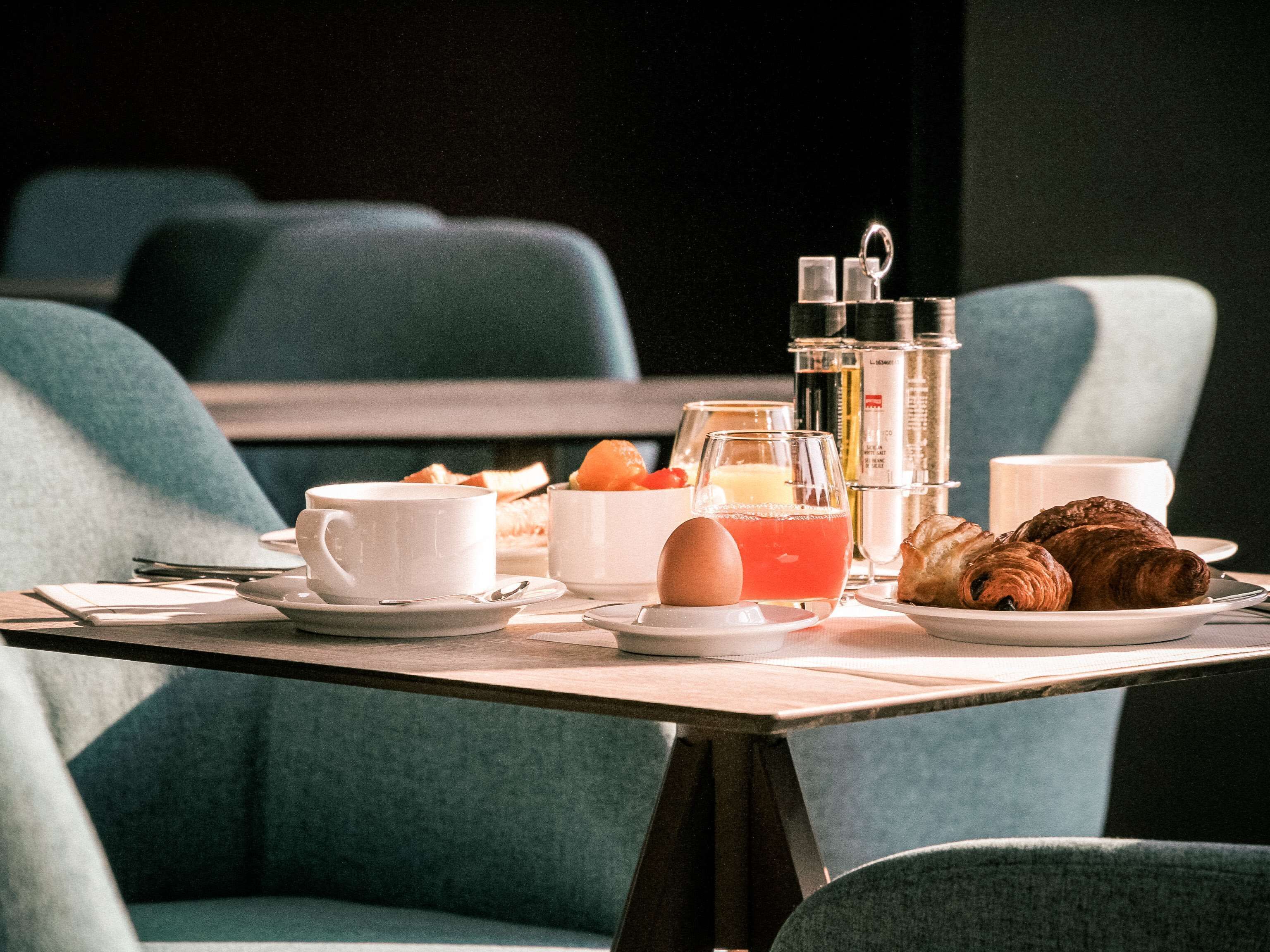 Preparación para el desayuno con una cafetera, croissants y un vaso de zumo de naranja sobre una mesa en una habitación con asientos tapizados en color turquesa.