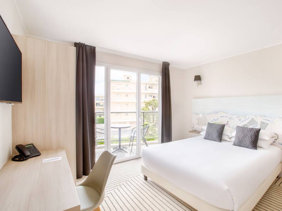 Habitación de hotel con cama, escritorio con teléfono, silla, ventana con cortinas y vista a edificios.