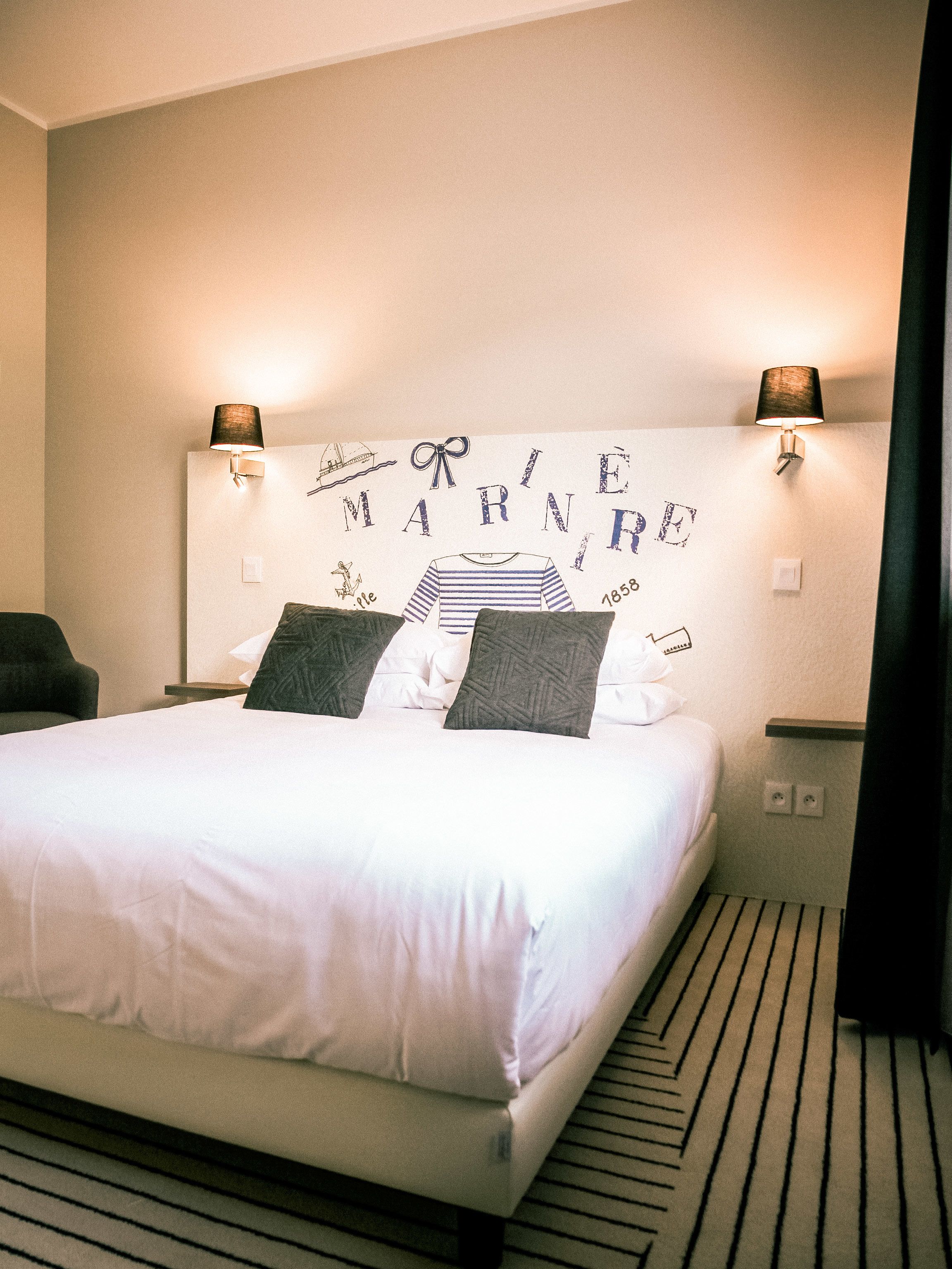 Una cama bien hecha con sábanas blancas y dos almohadas, un cabecero con la palabra “MARINE” y una ilustración de un barco, dos lámparas de pared y una alfombra a rayas.