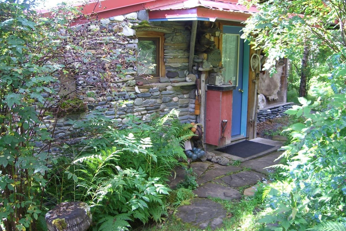 A rustic stone building with a red roof, a wooden door, and a small porch area.