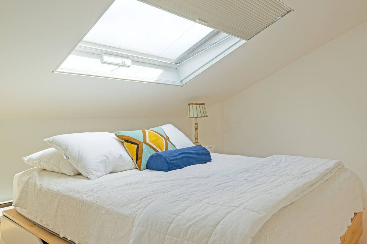 Een slaapkamer met dakraam, een bed met wit beddengoed, een lamp en een stopcontact.