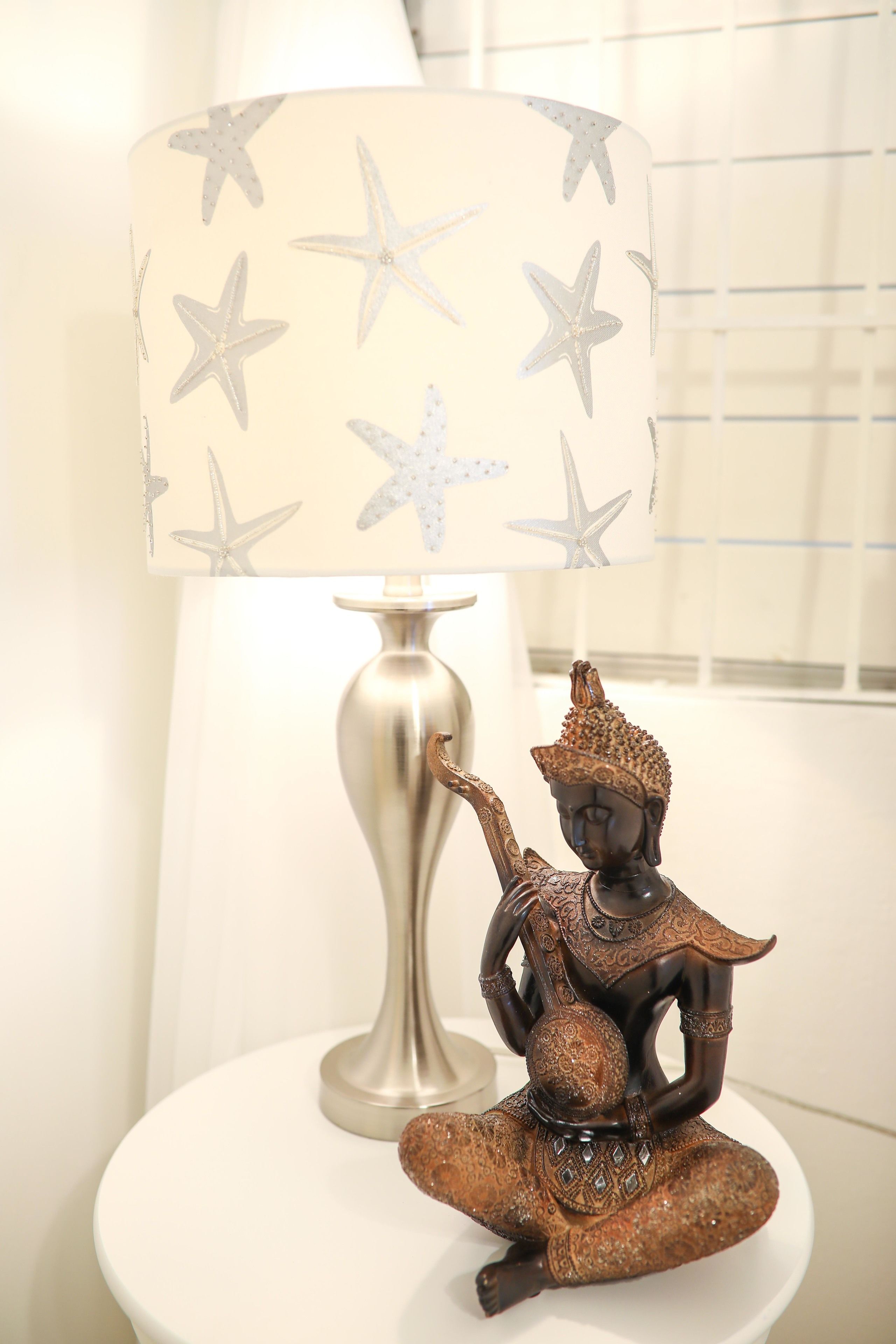 Una lámpara con pantalla estampada con motivos de estrellas y una figura decorativa de Buda sobre una mesa blanca.