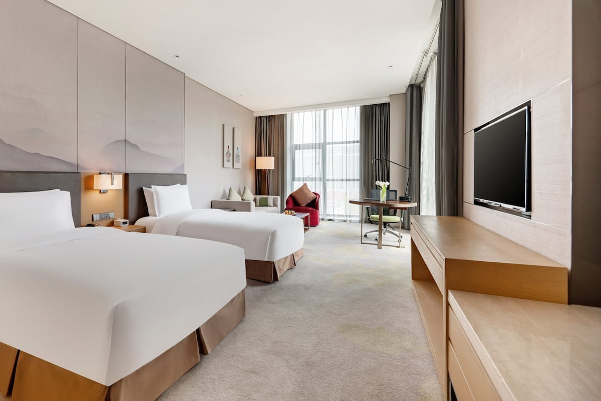 Une chambre d’hôtel avec deux lits, un bureau avec une chaise, une télévision et une grande fenêtre avec des rideaux.