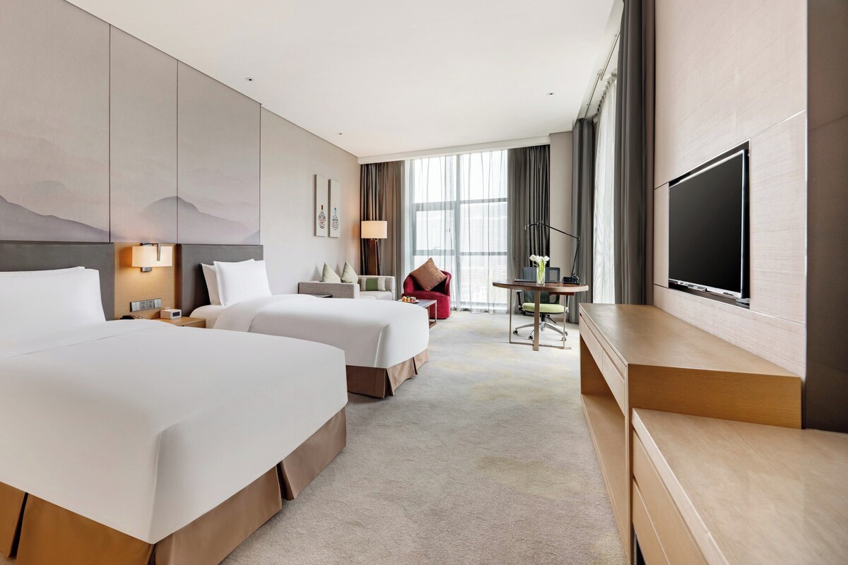 A hotel room with two beds, a desk with a chair, a TV, and a large window with curtains.