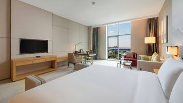 A hotel room with a large bed, a desk with a chair, a television, and a view of the city.