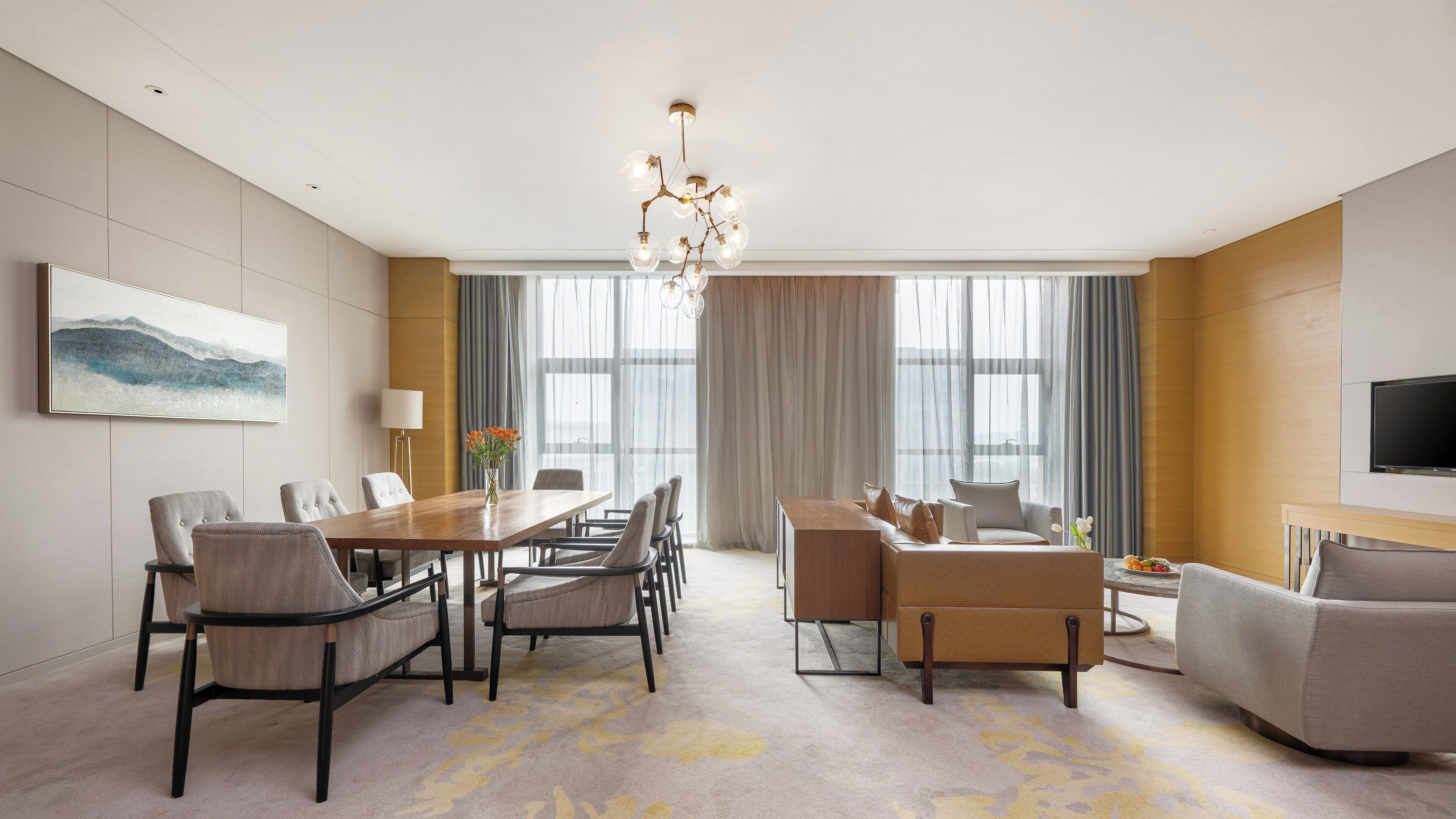 A modern hotel room with a dining area, a sofa set, a TV, and a large window with curtains.