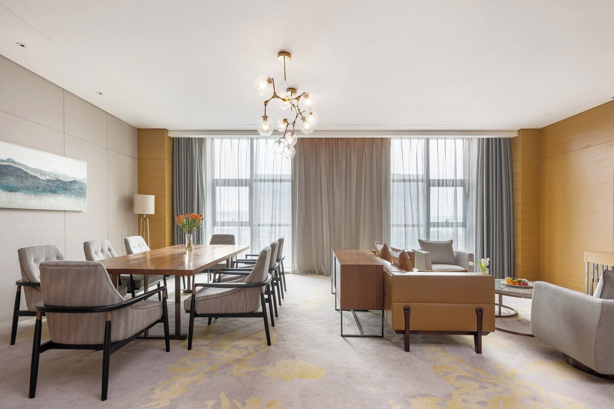A modern hotel room with a dining area, a sofa set, a TV, and a large window with curtains.