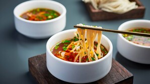 Eine Schüssel Nudelsuppe, mit Stäbchen, die Nudeln greifen, begleitet von einer kleinen Saucenschale und einer Nudelschale im Hintergrund.
