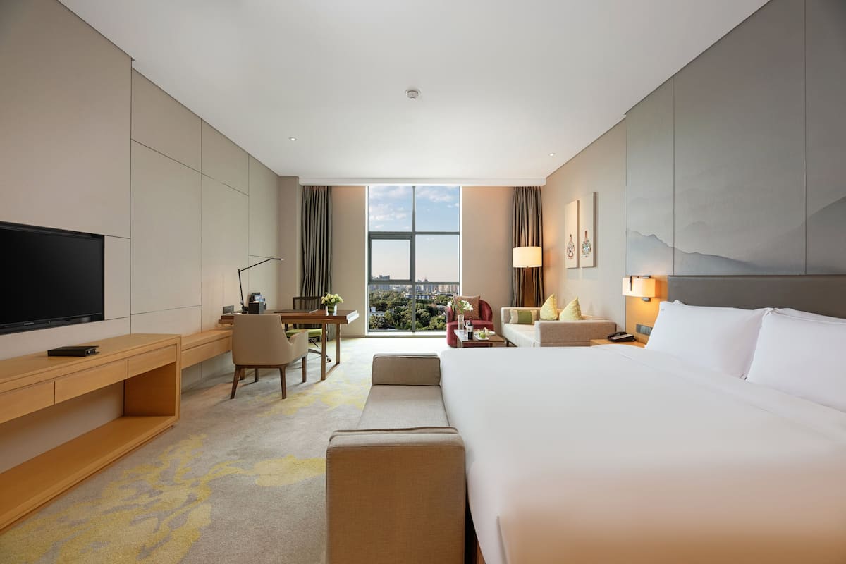 Une chambre d’hôtel avec un grand lit, un bureau avec une chaise, une télévision et une vue sur la ville grâce à de grandes fenêtres.