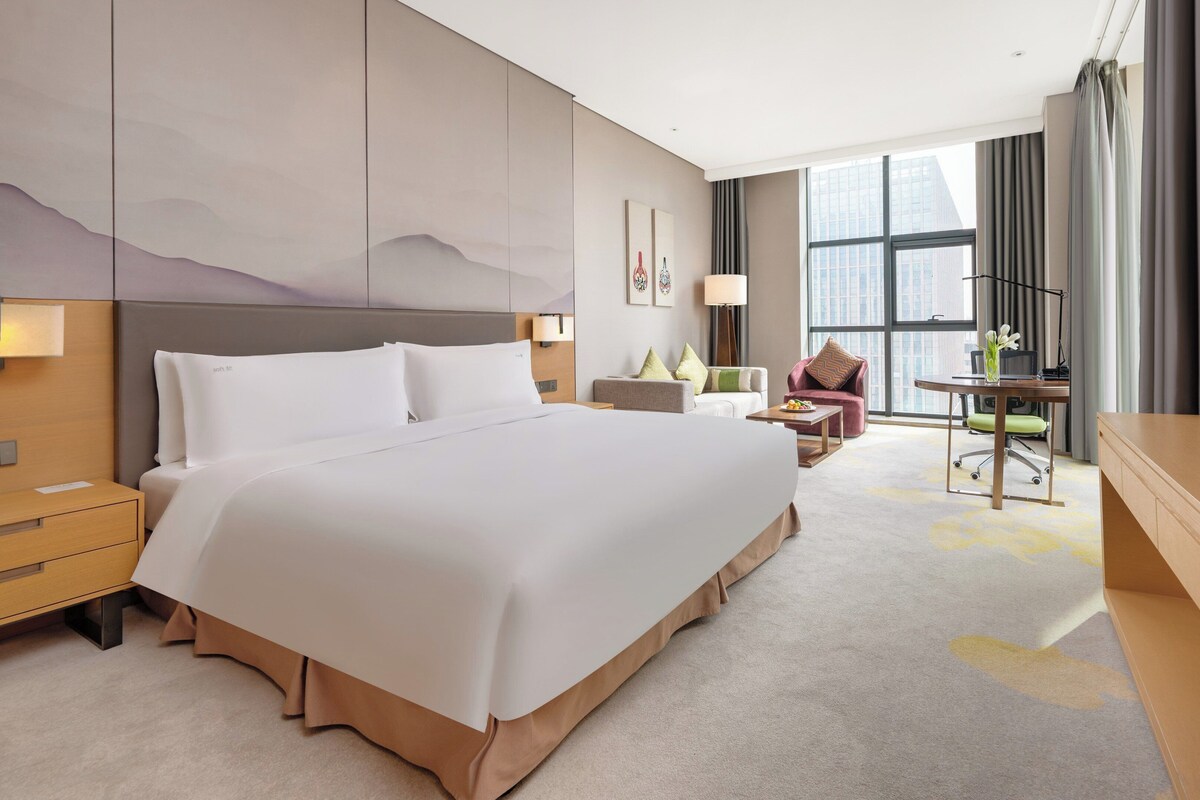 A hotel room with a large bed, a desk with a chair, a TV, and a window with curtains.