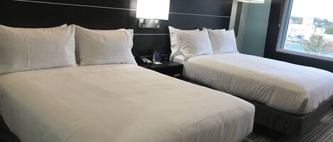 A hotel room with two beds, a nightstand, a lamp, and a window with a view.