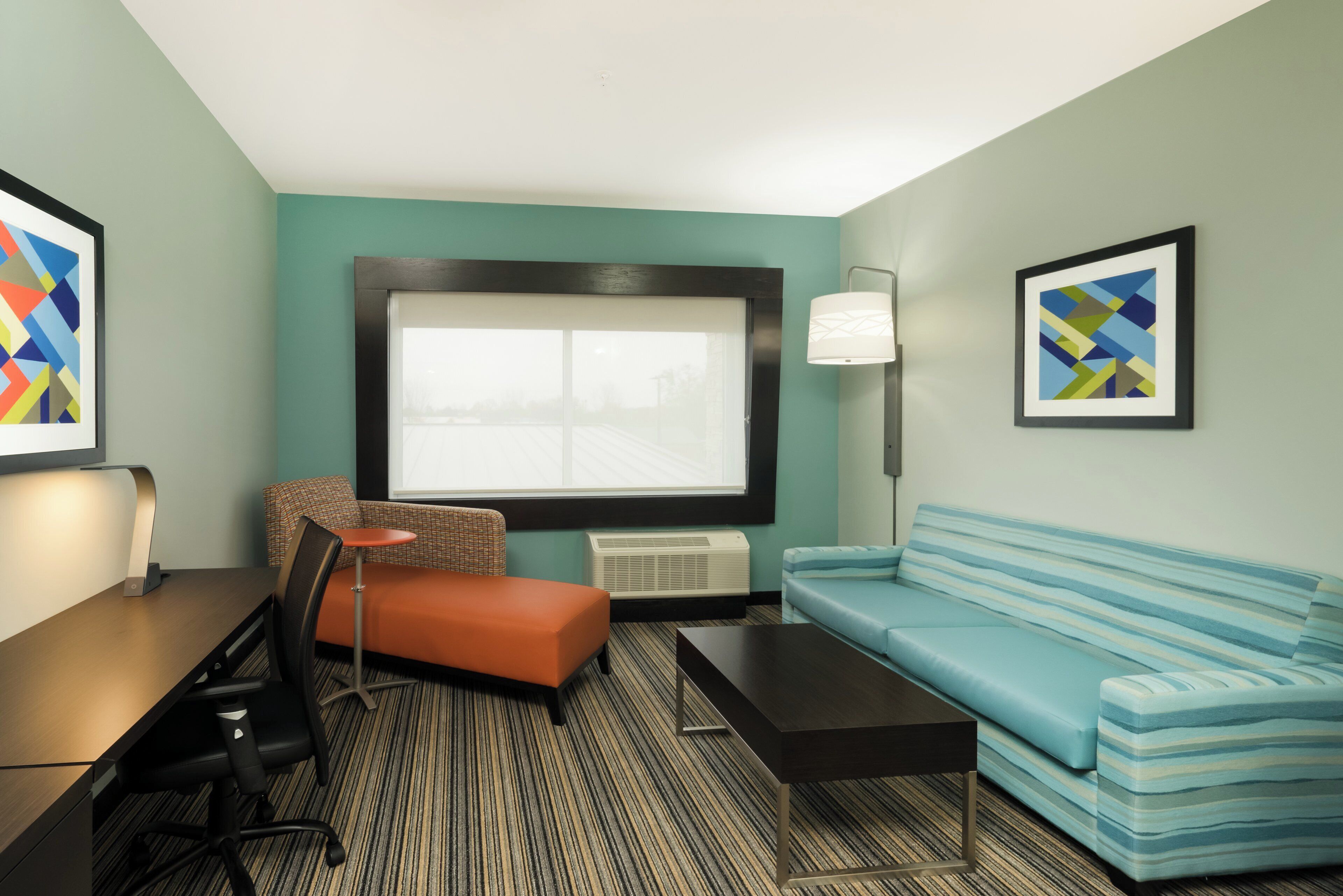 A hotel room with a large window, a sofa, a small table, a chair, a lamp, and artwork on the walls.